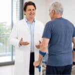 Priv.-Doz. DDr. Maximilian Kasparek, MSc spricht mit einem älteren Patienten, der mit zwei Unterarmstützen steht.