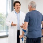 Priv.-Doz. DDr. Maximilian Kasparek, MSc speaks with an elderly patient who is standing with two forearm crutches.